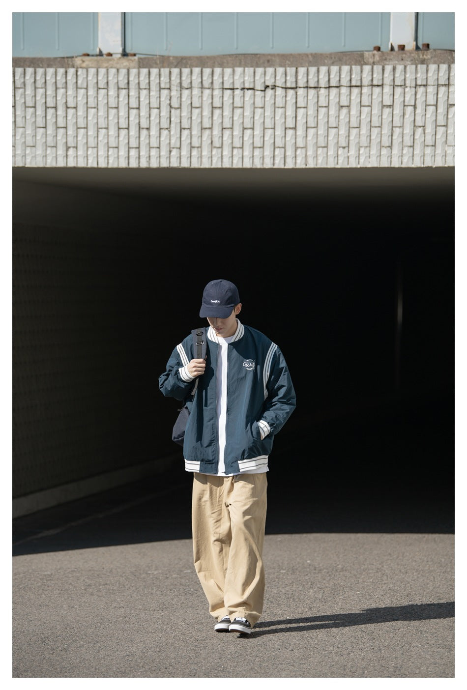 Full-length model wearing a dark navy zip jacket with white striped trim, beige trousers and a navy cap.
