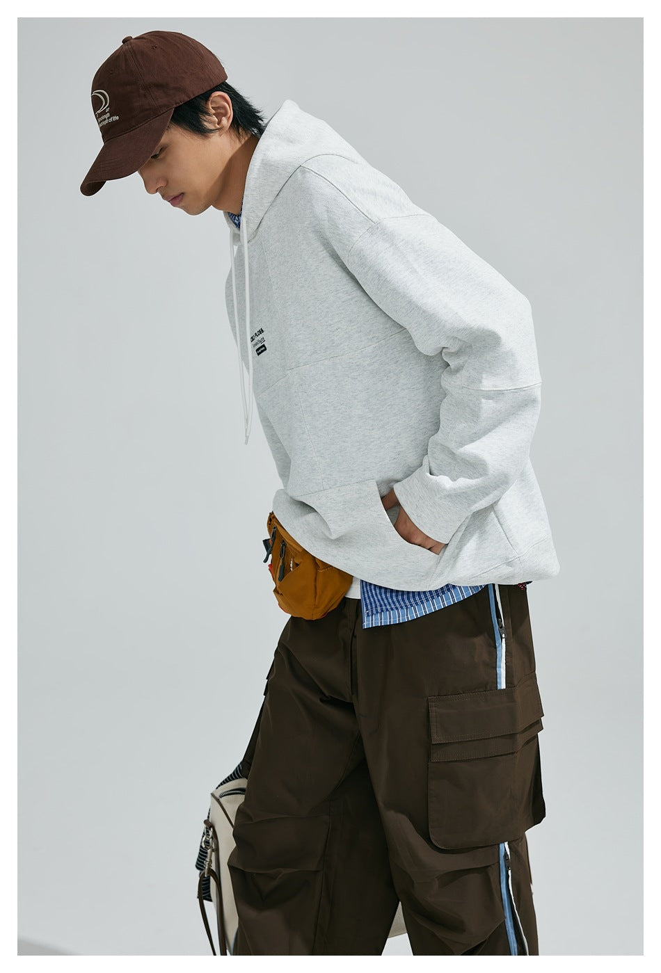 Model leaning forward in a light gray hooded sweatshirt with a front kangaroo pocket.