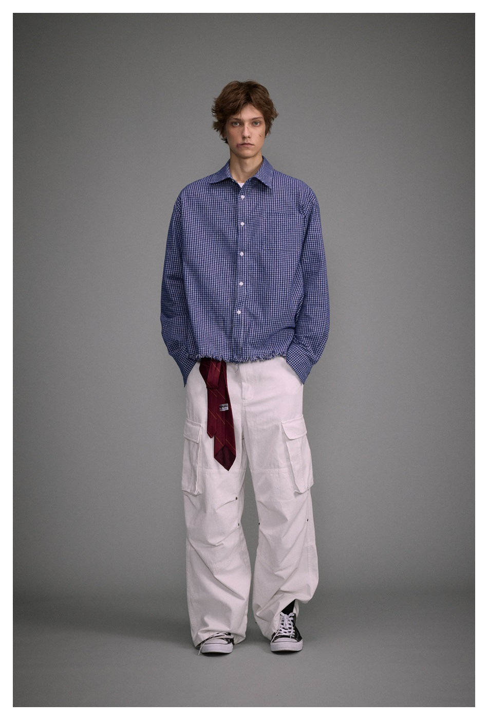 Model wearing a blue check button-up shirt with frayed hem, white cargo pants and a red tie, front view