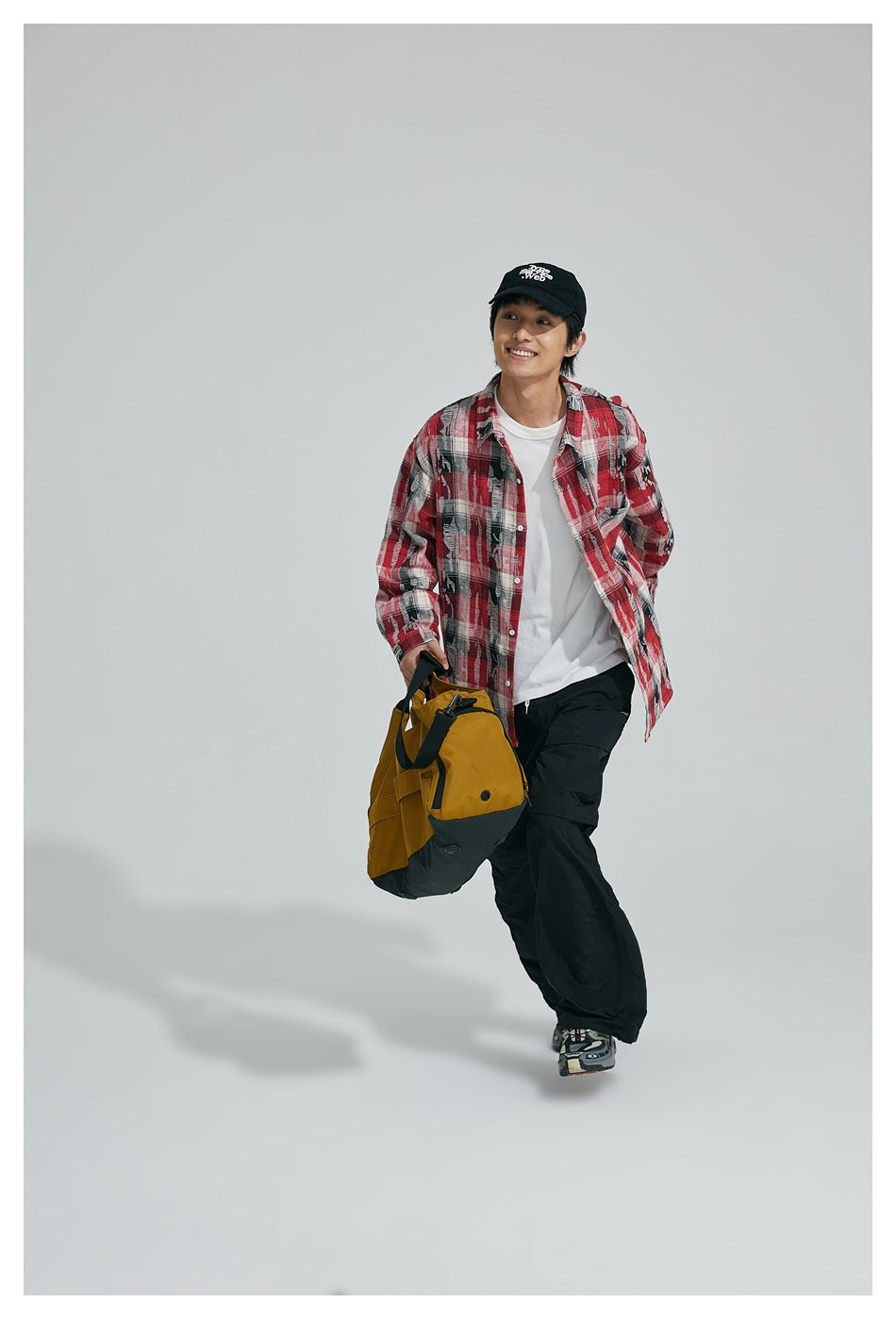 Model wearing a red, black, and white plaid button-up shirt with black pants and a cap, carrying a yellow bag.
