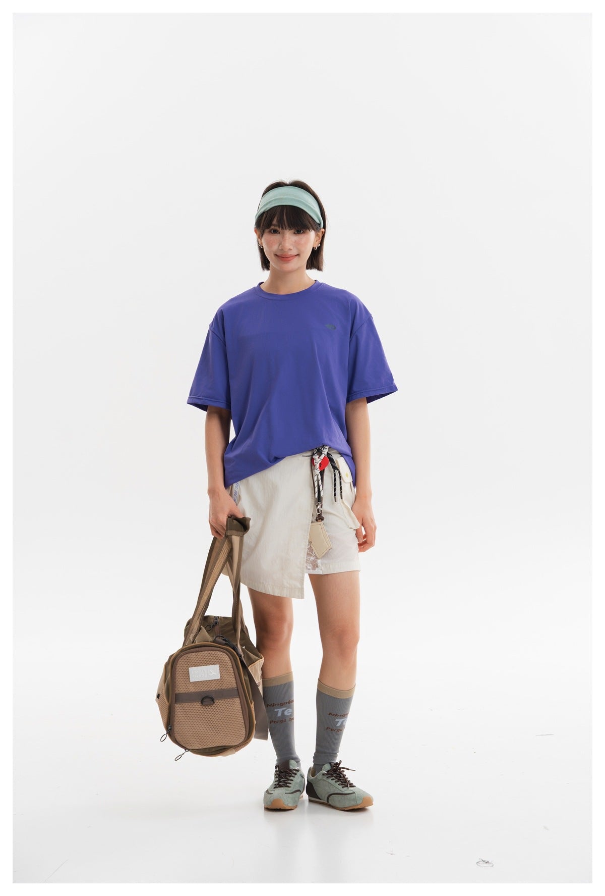 Front view of a model in an off-white utility skirt and loose purple T-shirt, holding a brown bag.