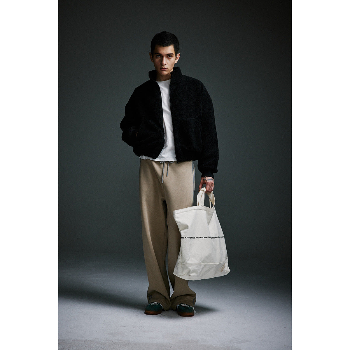 Full-length view of a model in a black fleece jacket and tan drawstring pants, carrying a white tote.