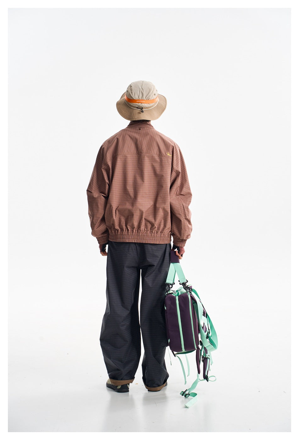 Rear view of a model wearing a rose checked jacket and loose dark checked trousers, holding a purple and mint bag.