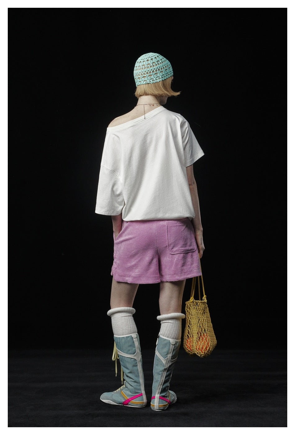 Back view of a model wearing a loose white T-shirt and lilac shorts, with a mint crocheted cap and a woven bag.