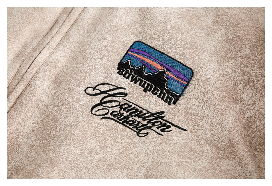 Close-up of embroidered mountain-and-sunset artwork and script on a beige mottled jacket.