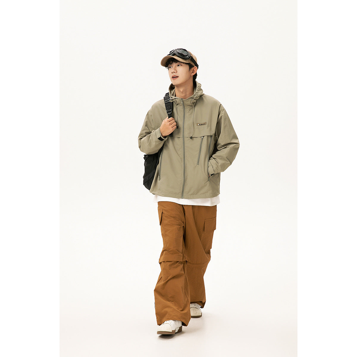 Full-length model wearing a sage-gray hooded jacket, brown cargo pants, white sneakers, and a cap with goggles.
