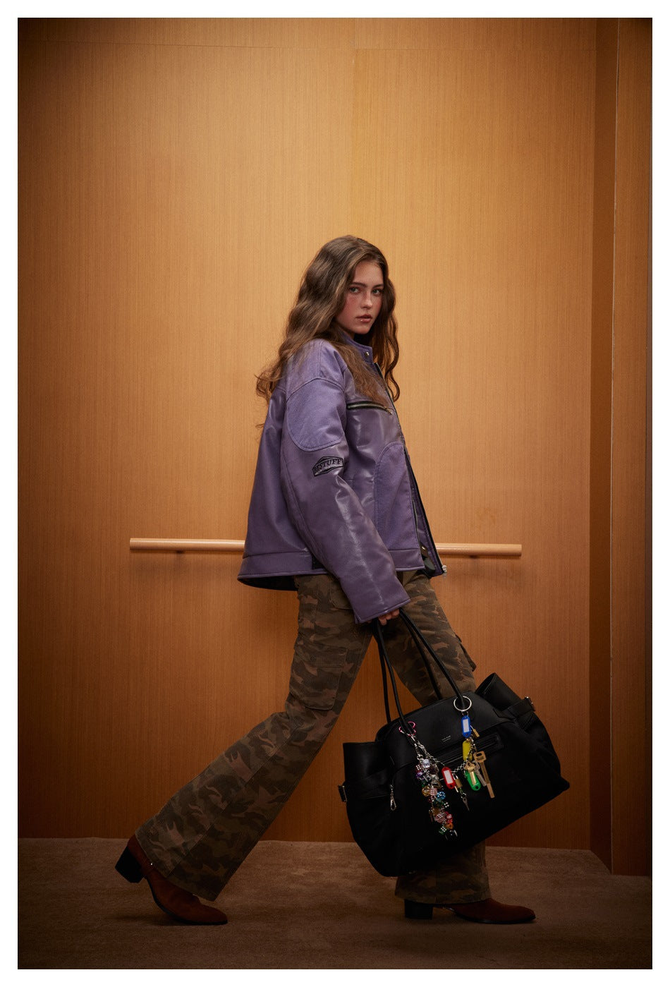 Model in purple leather-look racer jacket with camouflage cargo pants carrying a black tote bag