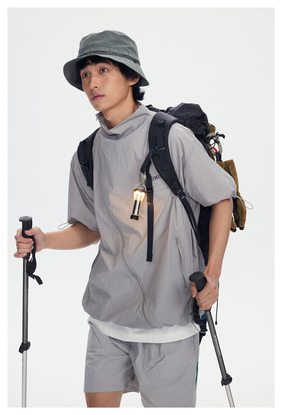 Close view of a model in a light gray zip jacket and bucket hat, carrying a backpack and hiking poles.