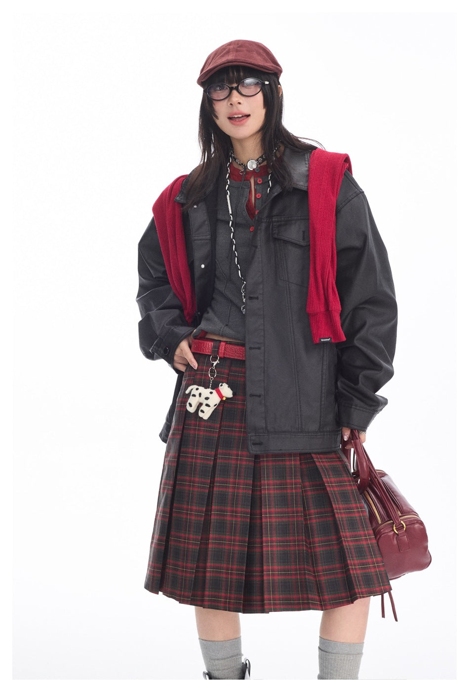 Model wearing a charcoal jacket over a gray top, red-and-black plaid pleated skirt, and red knit layer.