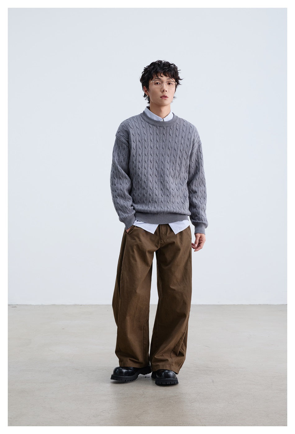 Front view of a model in a gray cable-knit crewneck layered over a striped shirt with brown trousers.
