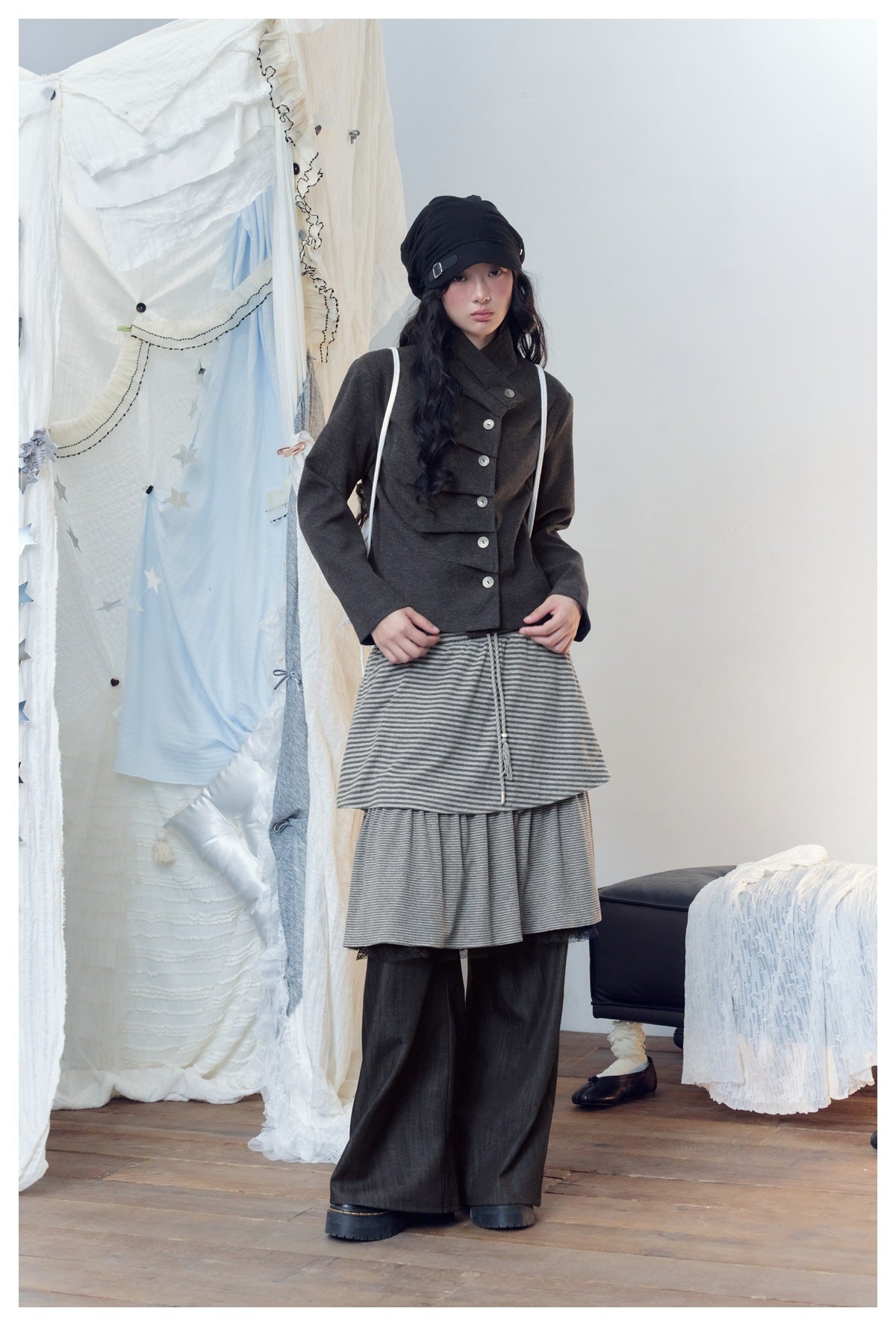 Model facing front wearing wide-leg pants with a gray striped tiered skirt layer, a gray jacket and black cap