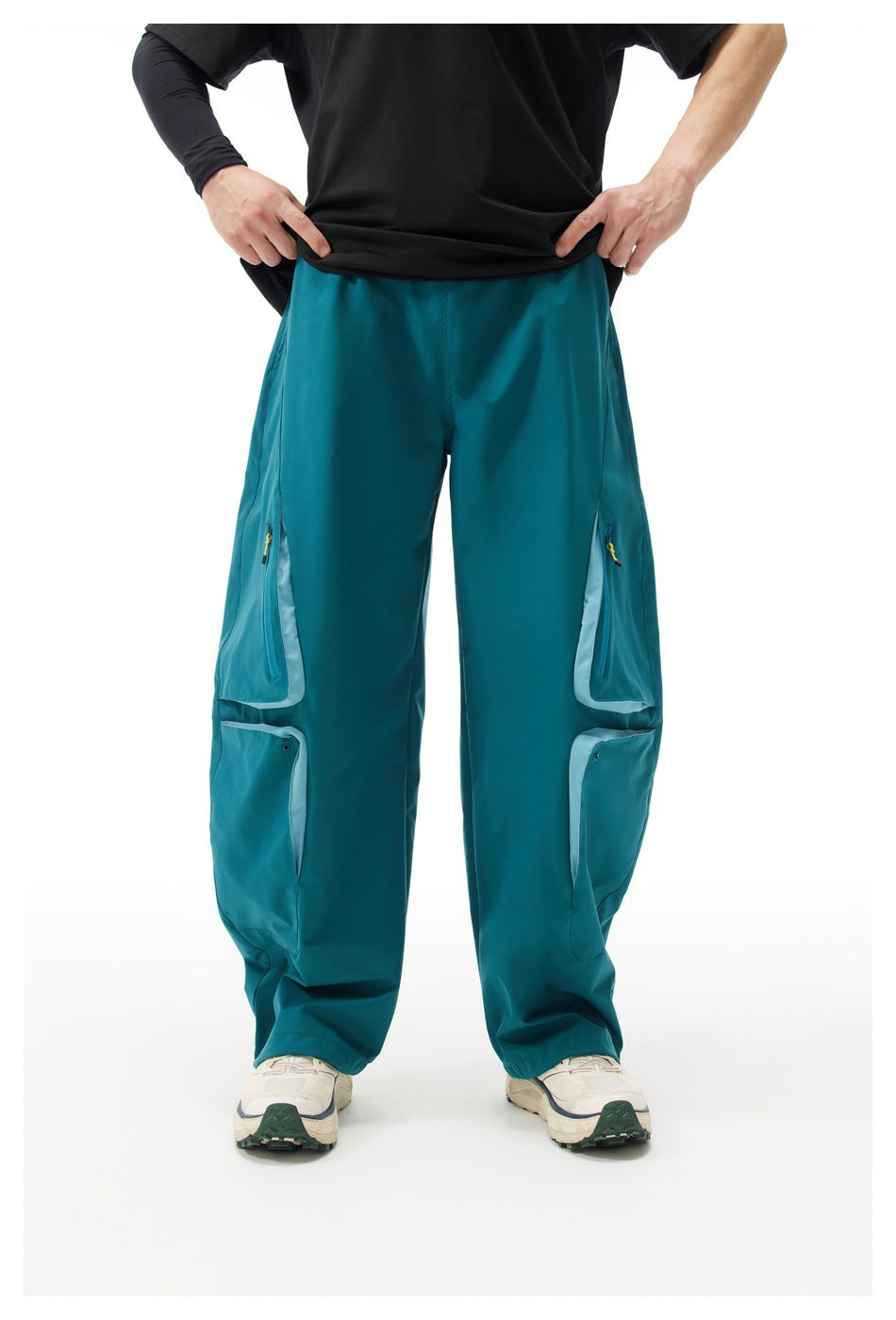 Front view of a model wearing loose teal cargo pants with paired thigh pockets, pale trim, and yellow zipper pulls.