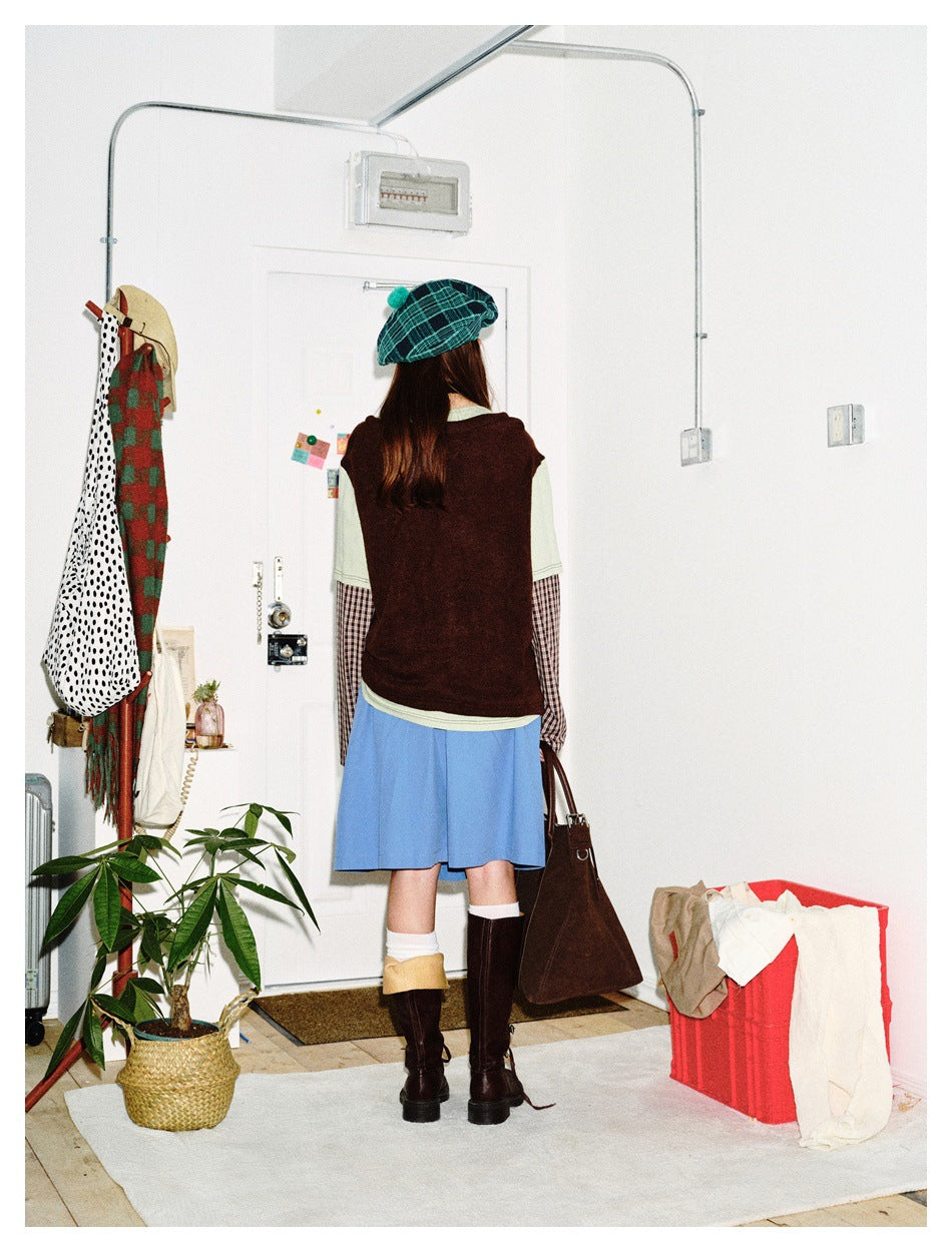 Rear view of a model wearing a burgundy top over sage and checked sleeves, a blue skirt, and a green plaid beret.