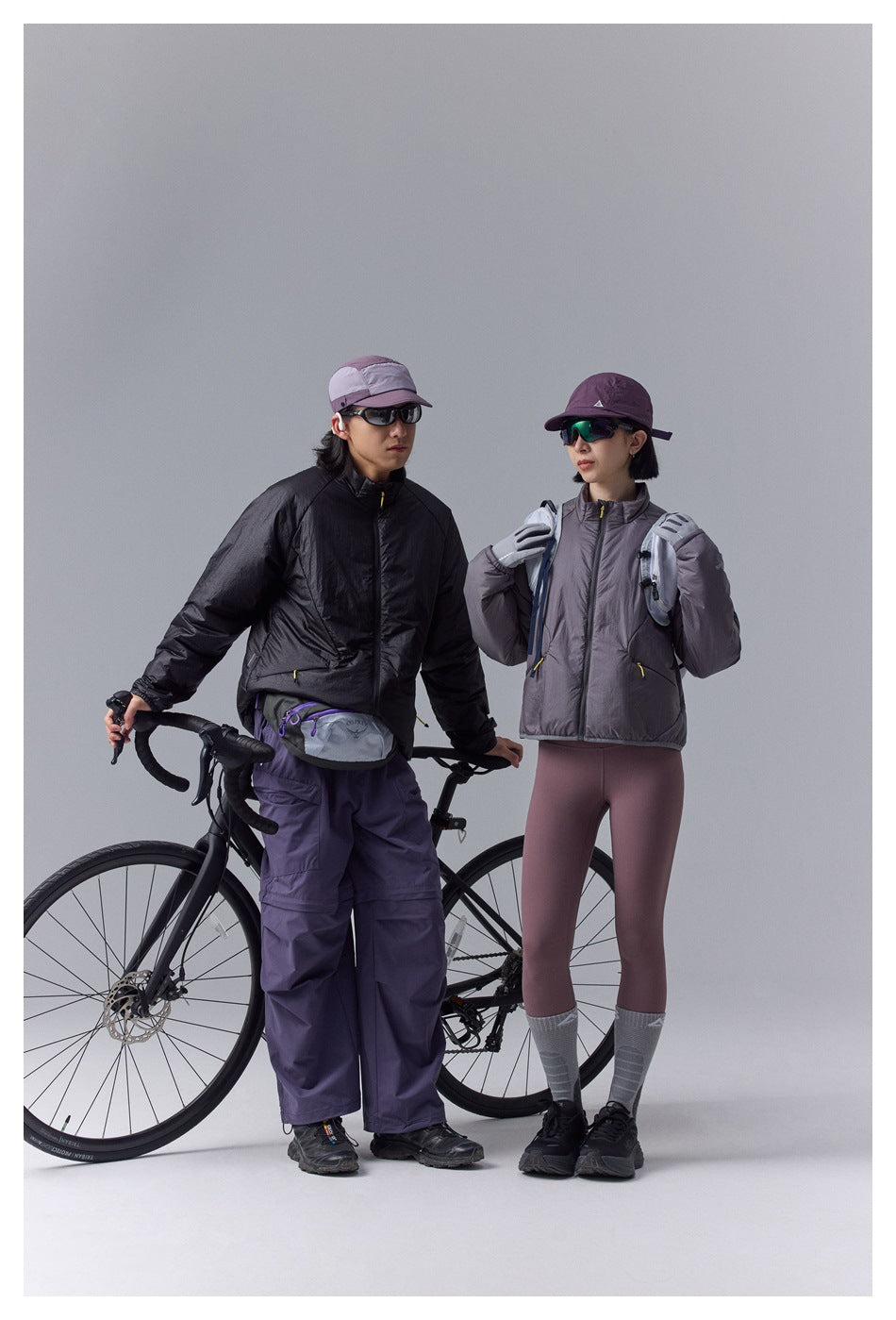 Two cyclists wear black and gray zip-front jackets with purple outdoor pants beside a road bike.