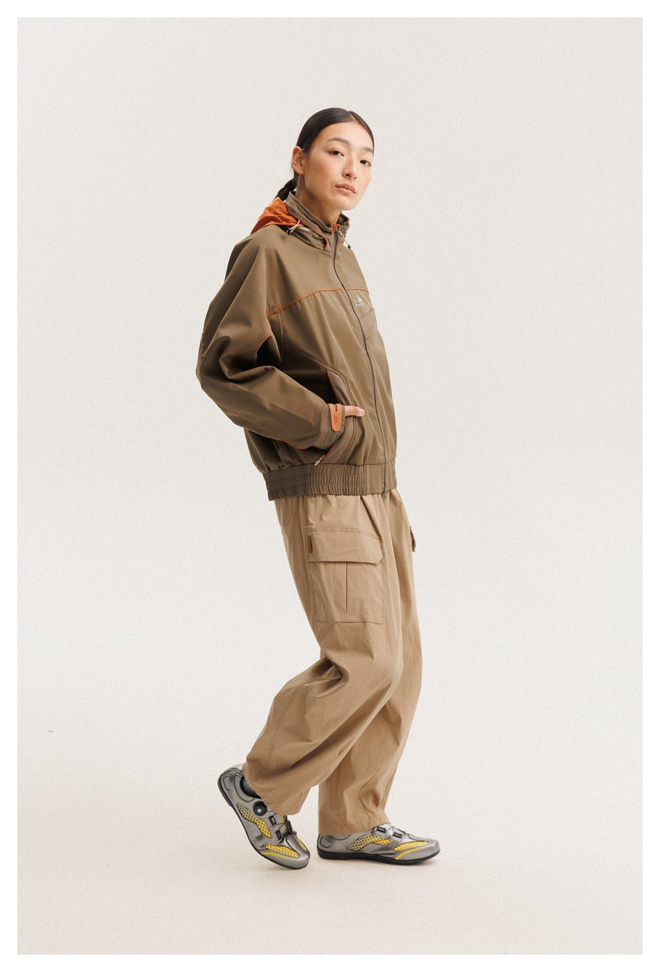 Model in a brown hooded jacket with orange trim and tan cargo pants, posing in a three-quarter side view.