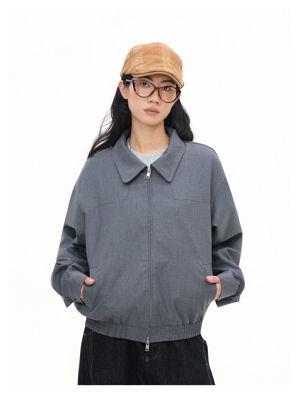 Model wearing zipped gray collared jacket with hands in pockets, dark jeans and tan cap