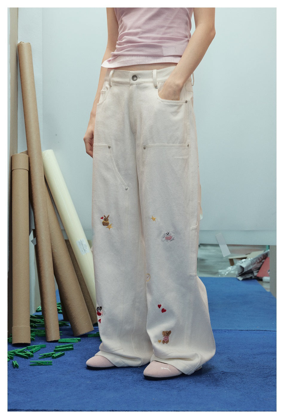 Angled front view of a model wearing cream wide-leg trousers with utility pockets and small embroidered motifs.