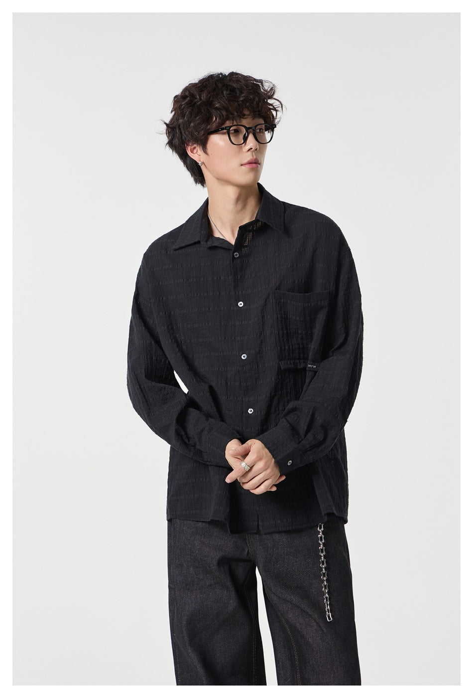 Model wearing a black textured button-up shirt with a chest pocket and dark jeans, shown from the front.