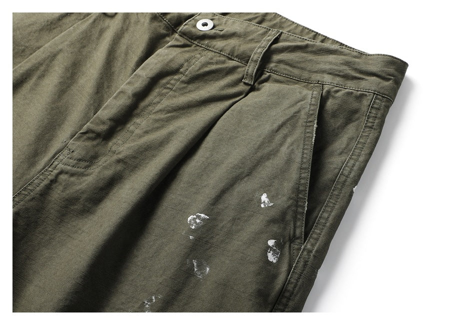 Close-up of olive pants showing a slanted pocket, waistband button, and scattered white distressed marks.