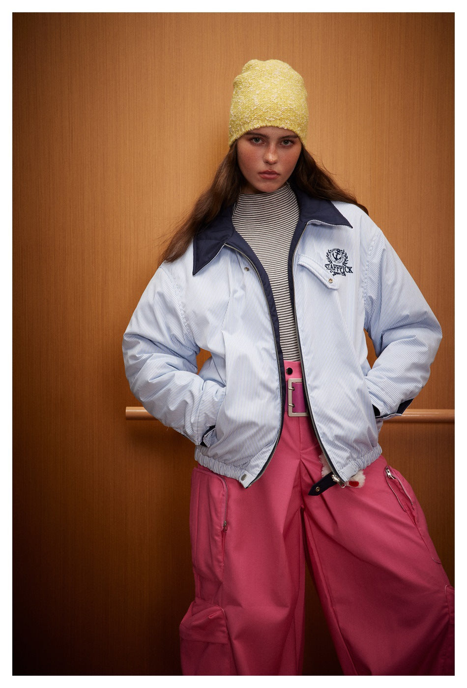 Model wearing a light blue pinstriped jacket with navy collar, yellow beanie and pink trousers.