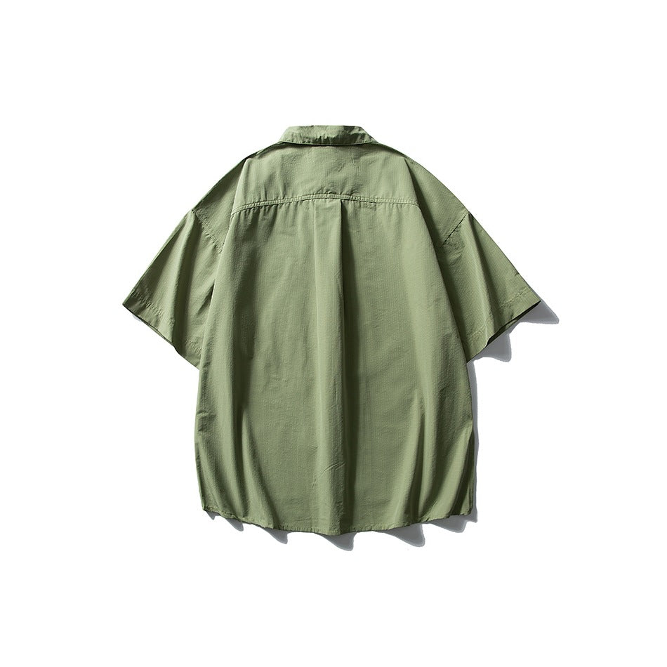 Back view of a sage green short-sleeve collared shirt with a center pleat and curved hem, displayed against white.