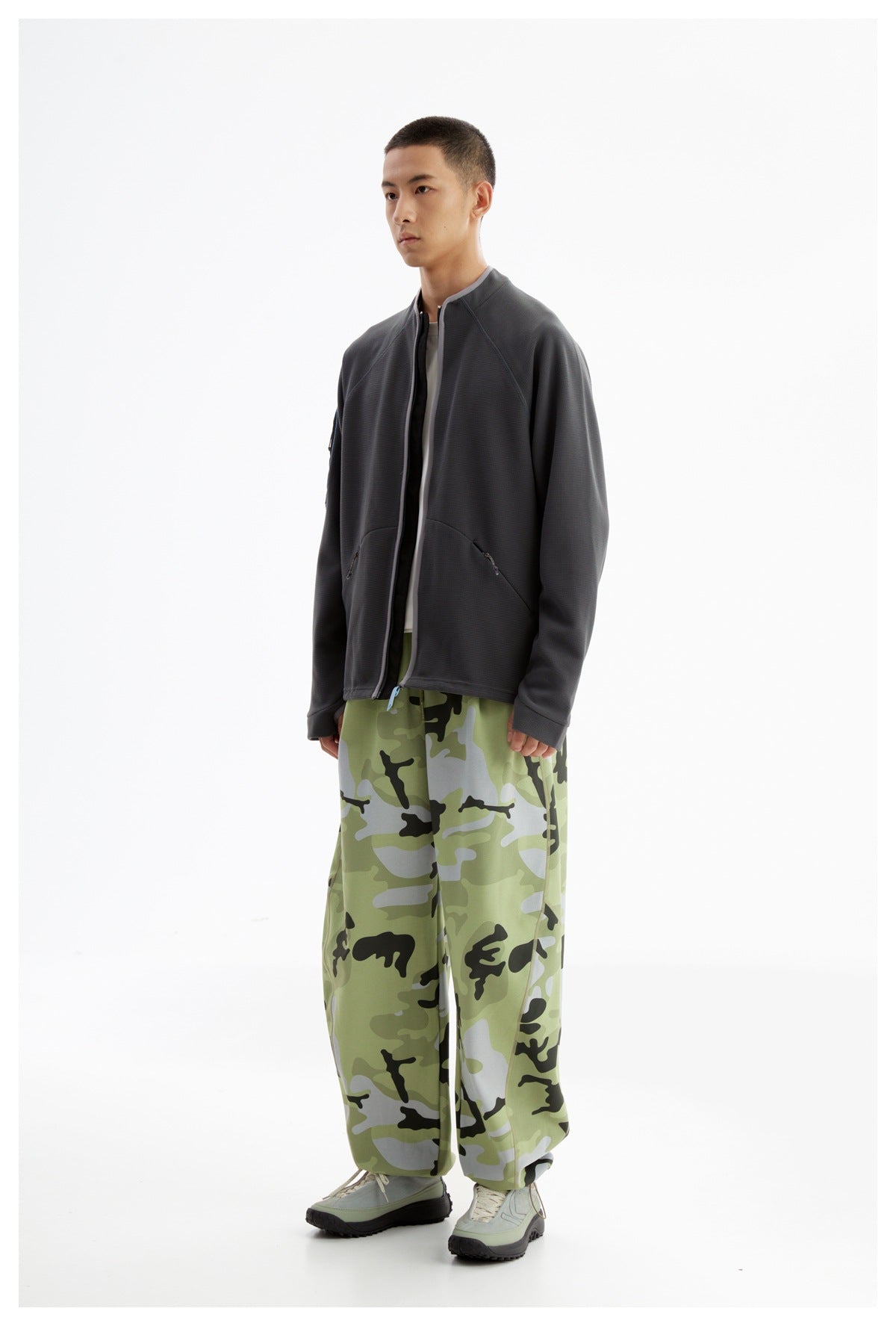 Model wearing a charcoal zip jacket with loose green camouflage pants, shown full length from a three-quarter angle.