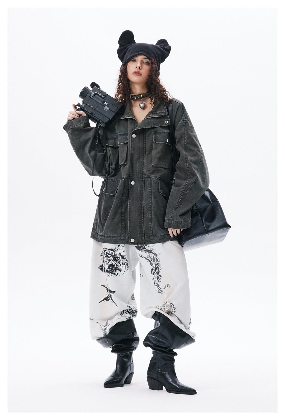 Model wearing an oversized charcoal jacket and white graphic trousers with black boots and a black ear-shaped beanie.