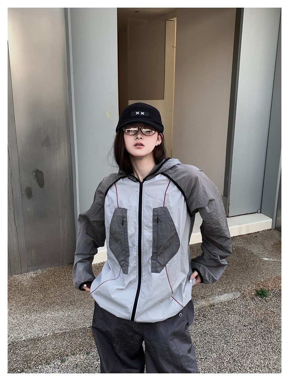 Model wearing a light gray hooded zip jacket with dark gray sleeves and pockets, red piping, and dark trousers.