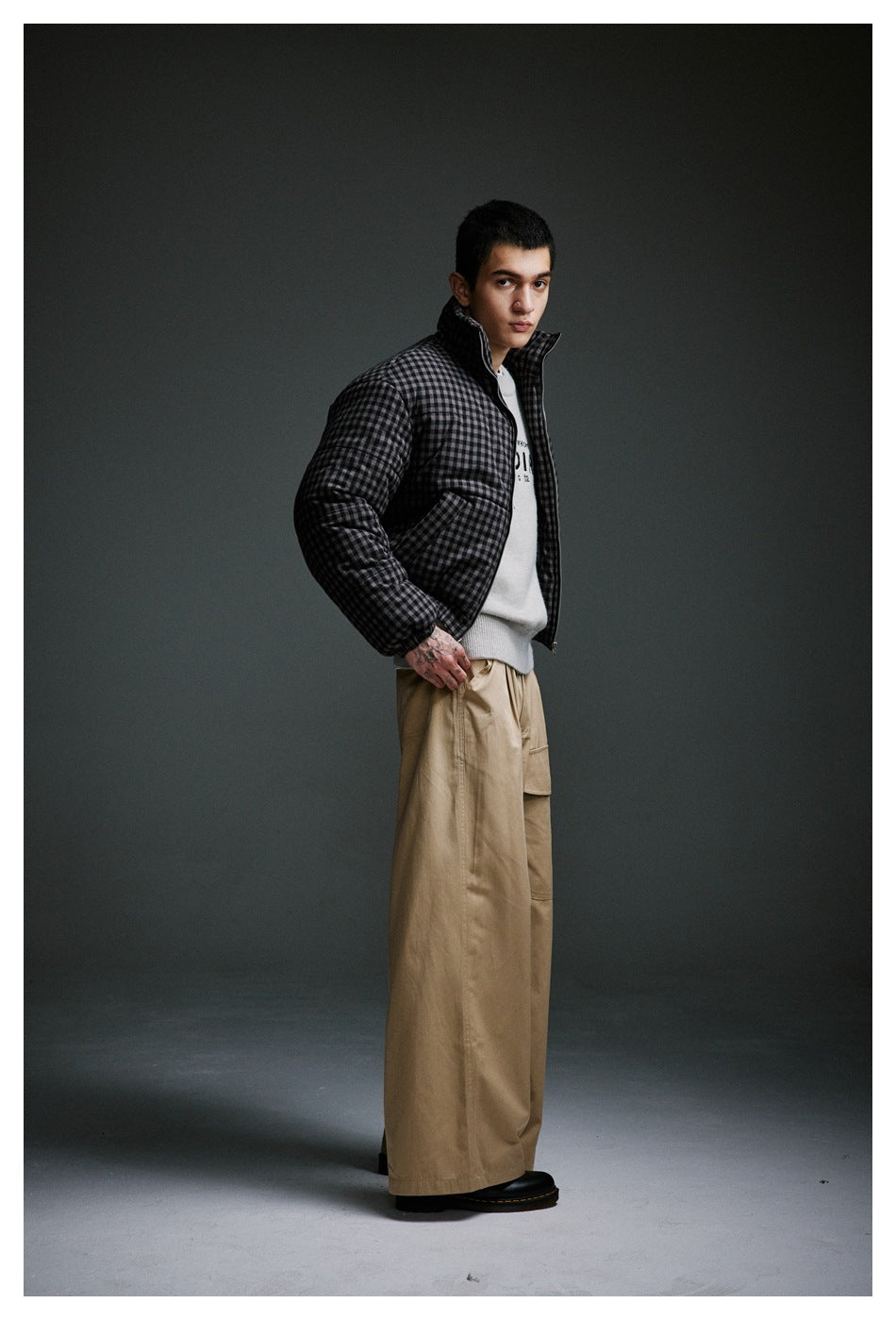 Model in a charcoal checked padded jacket, white sweater, and wide tan trousers, shown at an angle.