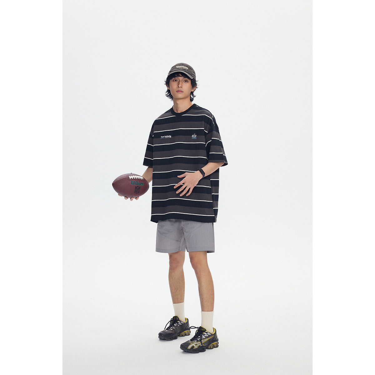 Model wearing an oversized black-and-charcoal striped T-shirt with light gray shorts, holding a football.