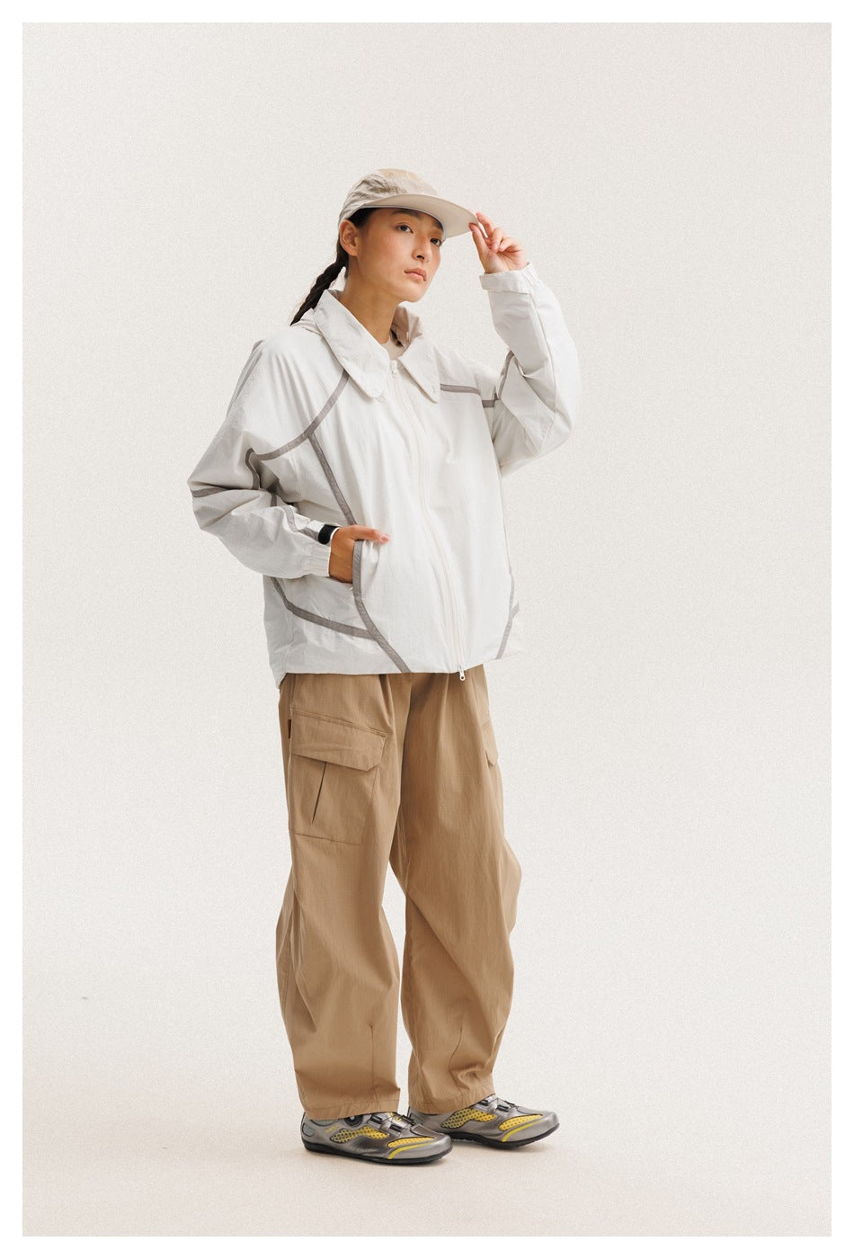 Model wearing a white zip-front jacket with gray piping and tan cargo pants, posing with one hand on a cap brim.