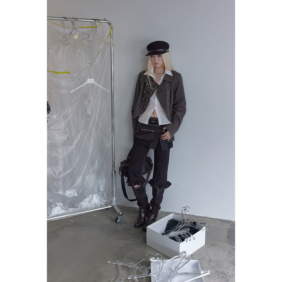 Full-length model in a charcoal asymmetrical jacket, white shirt, black ruffled trousers, boots and a black cap.