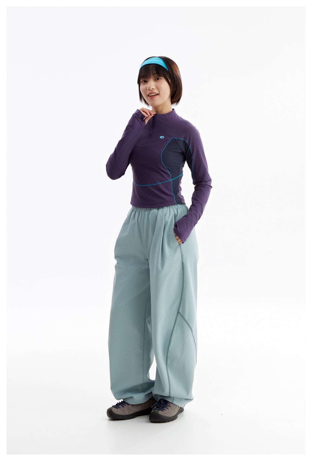 Model wearing a purple quarter-zip top with teal trim and loose pale blue pants, shown nearly full-length.