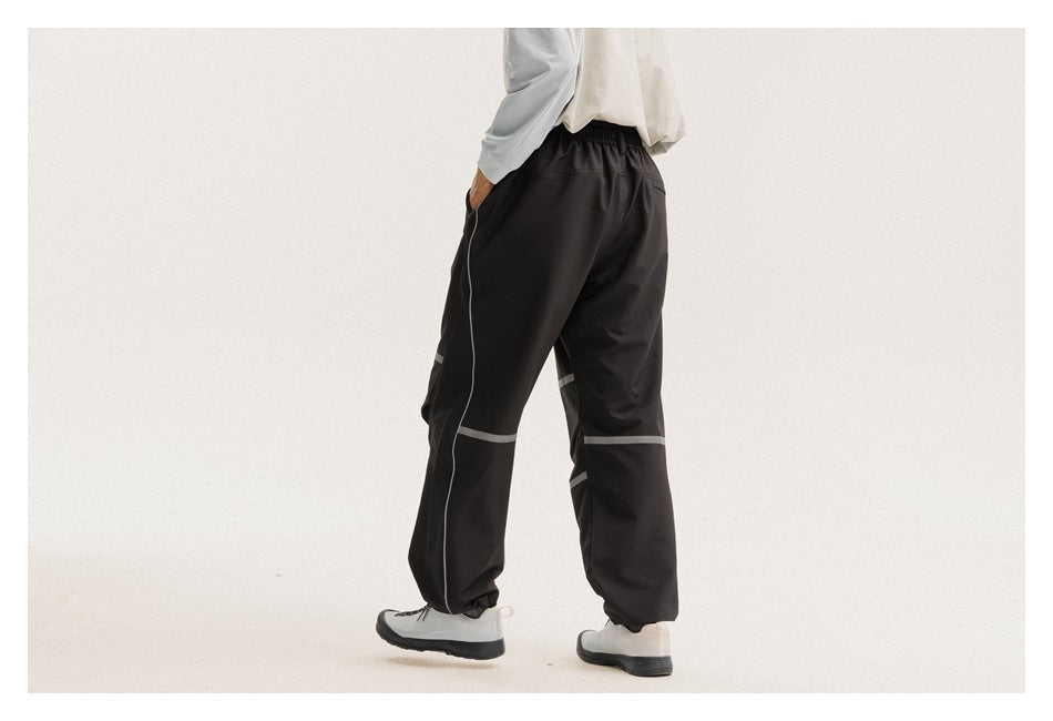 Rear view of a model wearing loose black pants with an elastic waist and gray bands across the legs.