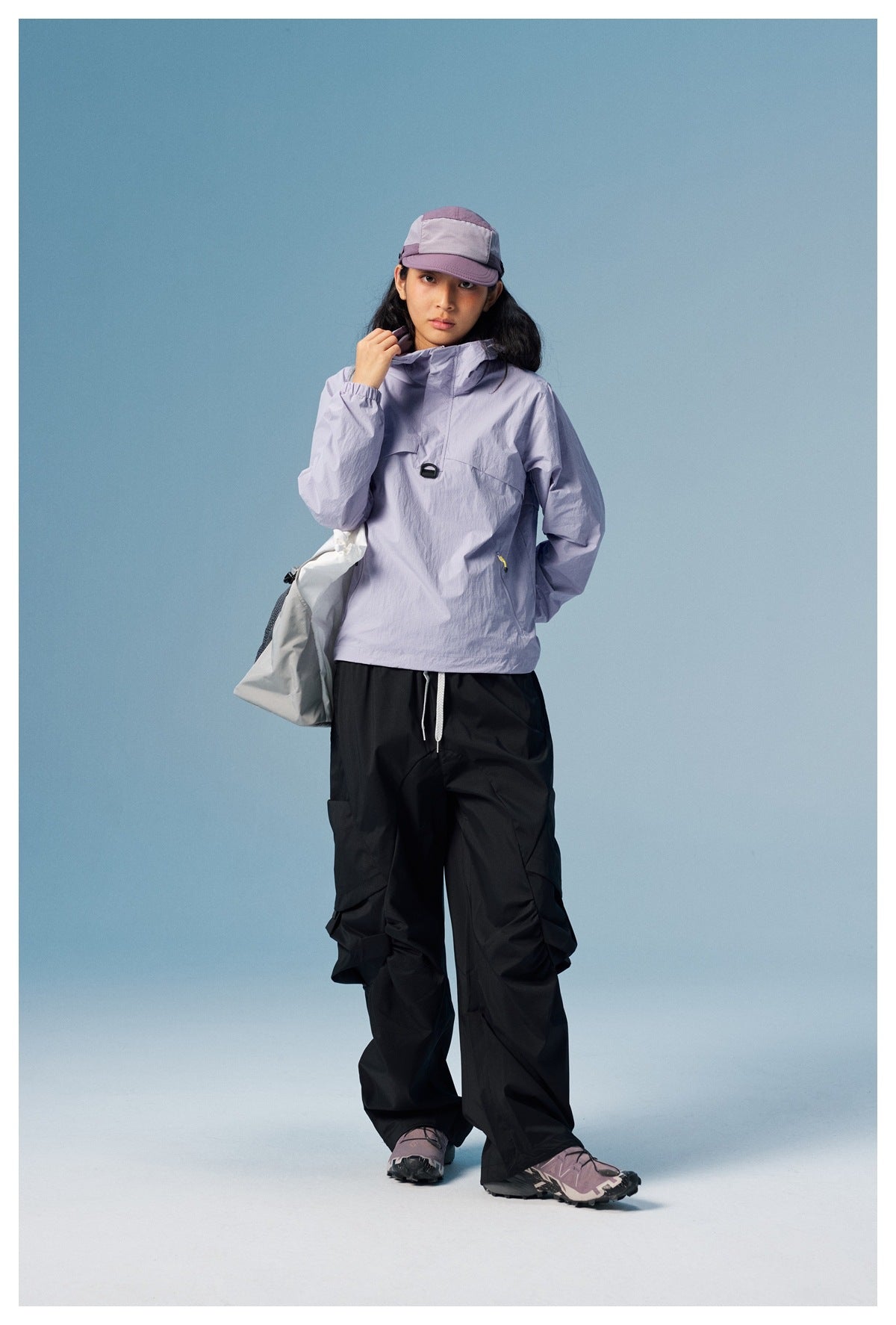 Model wearing a lavender hooded pullover with black cargo trousers and carrying a gray bag.