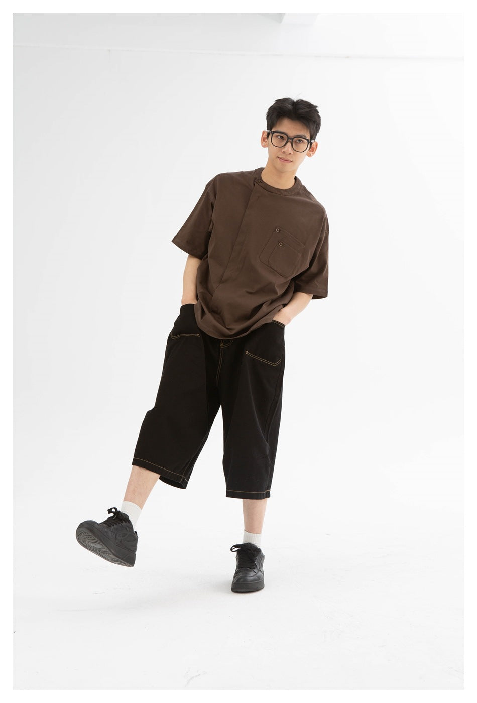 Model wearing a brown pocket tee and black wide-leg shorts with contrast stitching, posing with one leg raised.