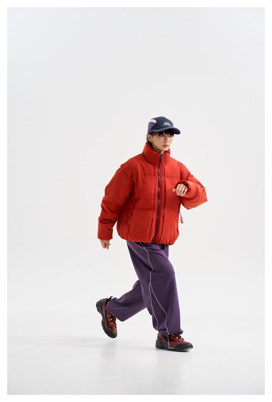 Model walking in a red-orange high-collar puffer jacket, purple trousers, and a navy cap.