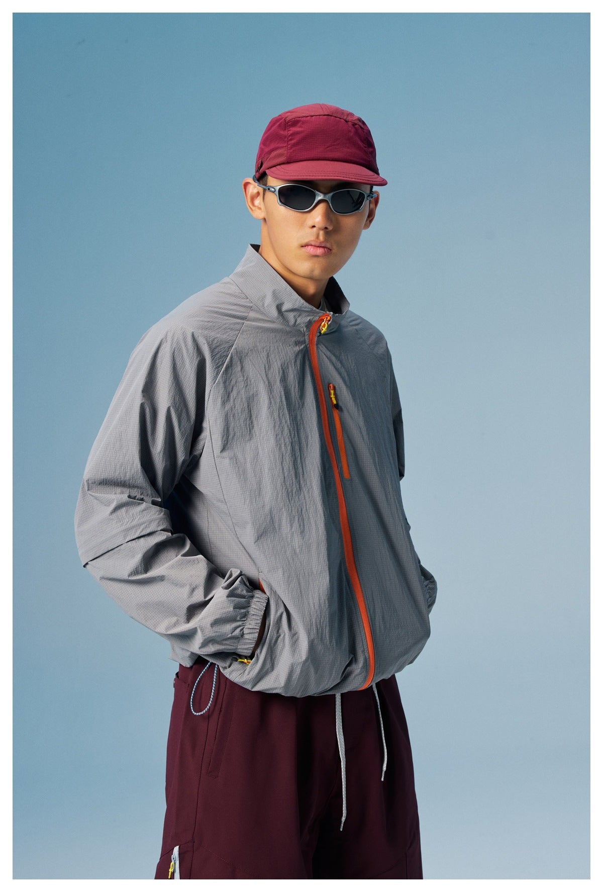 Close three-quarter view of a model wearing a gray high-collar jacket with orange zips and burgundy pants.