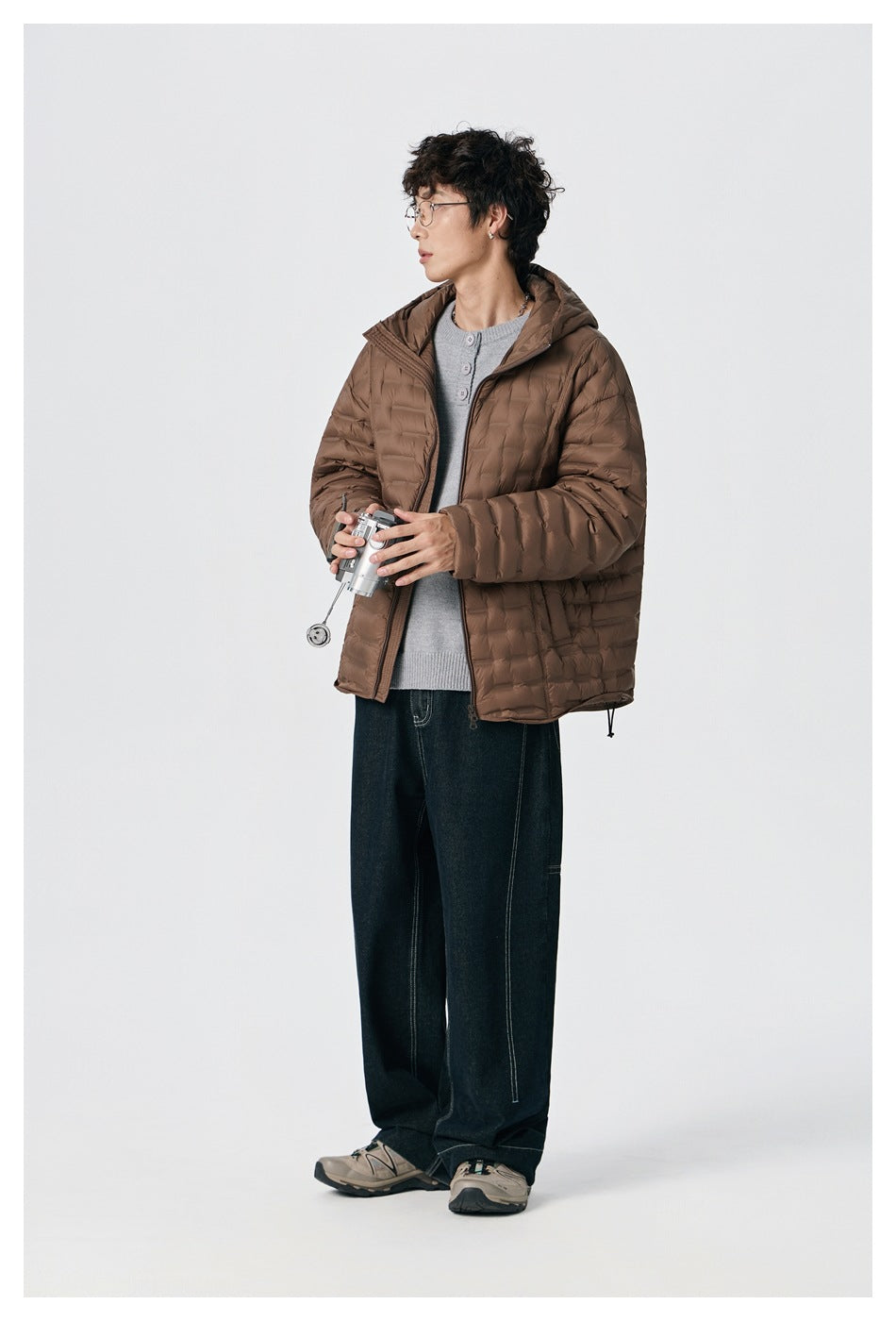 Model wearing a brown hooded quilted jacket over a gray cardigan and dark jeans, holding a small metal object.