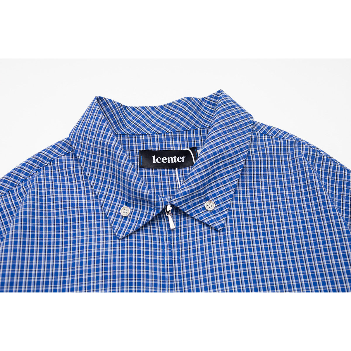 Close-up of a blue plaid shirt collar with two buttons, a center zipper, and a sewn black neck label.