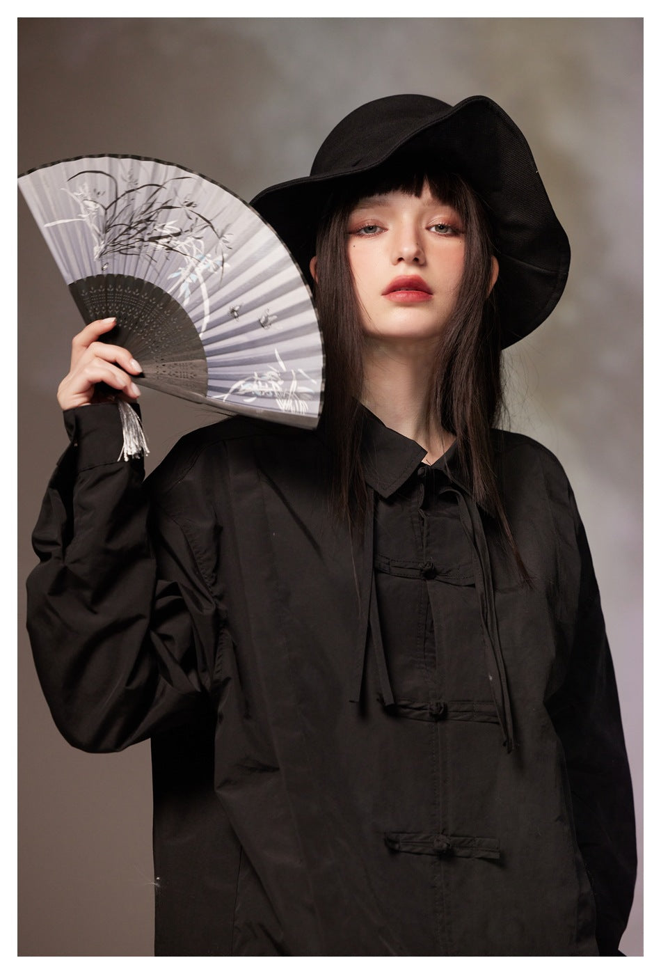 Model wearing a black collared shirt with frog closures and a wide-brim hat, holding an open illustrated fan.