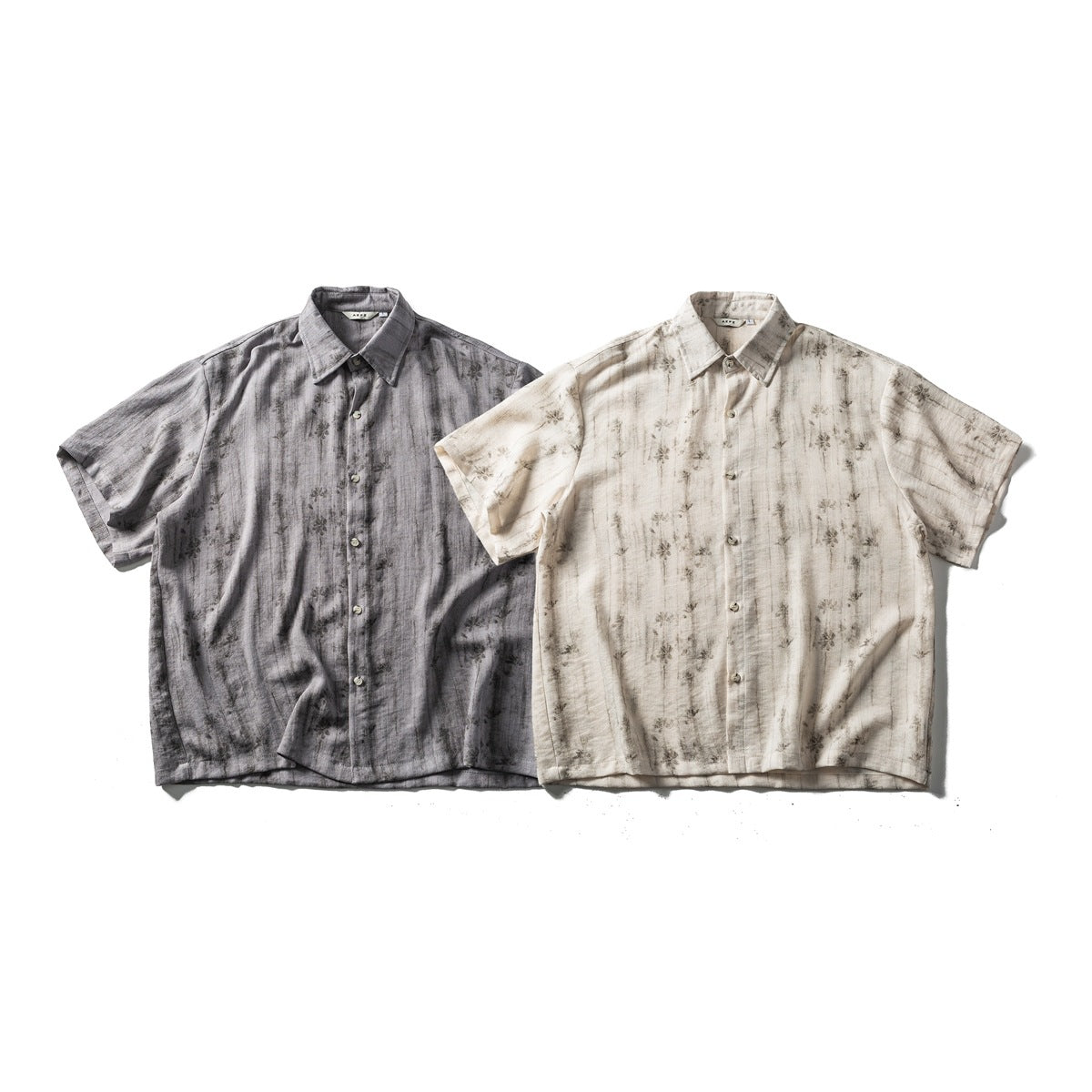 Two short-sleeve button-up shirts laid flat: one charcoal gray and one cream, both with dark mottled patterns.