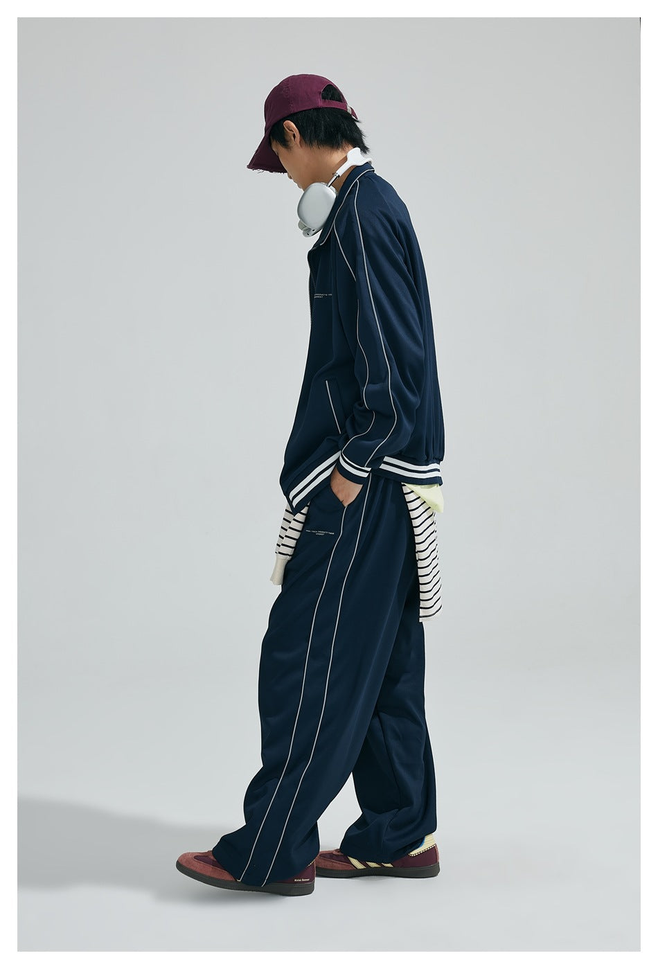 Side view of model in navy zip jacket with sleeve piping and striped cuffs, matching piped pants, maroon cap and sneakers