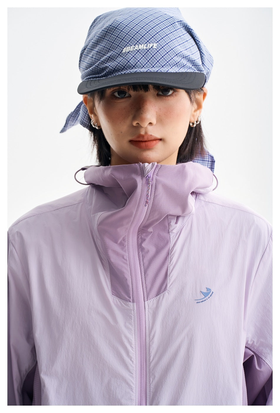 Close view of the lilac jacket's high hood, front zipper and small chest emblem, with a blue plaid cap.