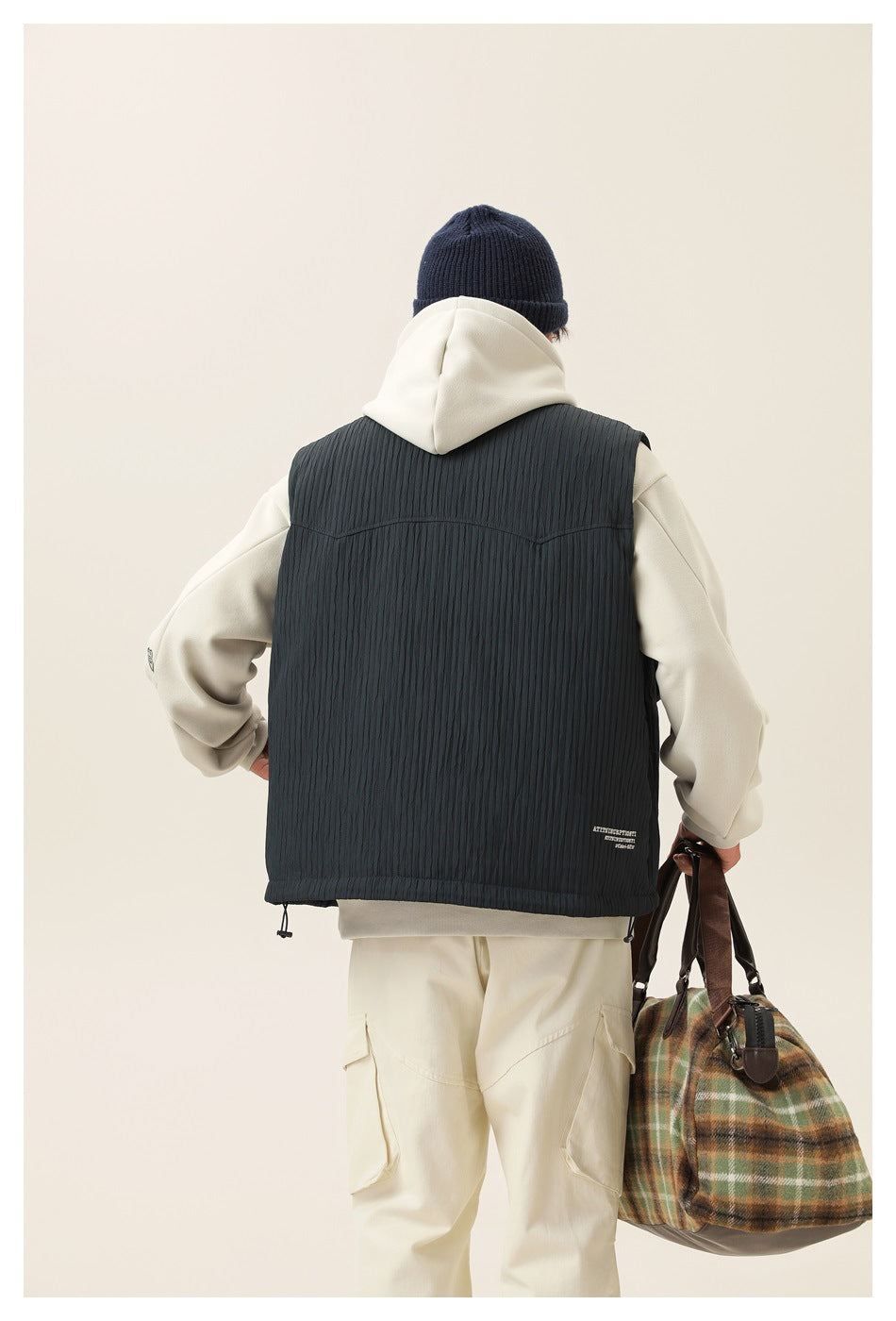 Back view of a model wearing a dark charcoal quilted vest over an ivory hoodie and cargo pants.