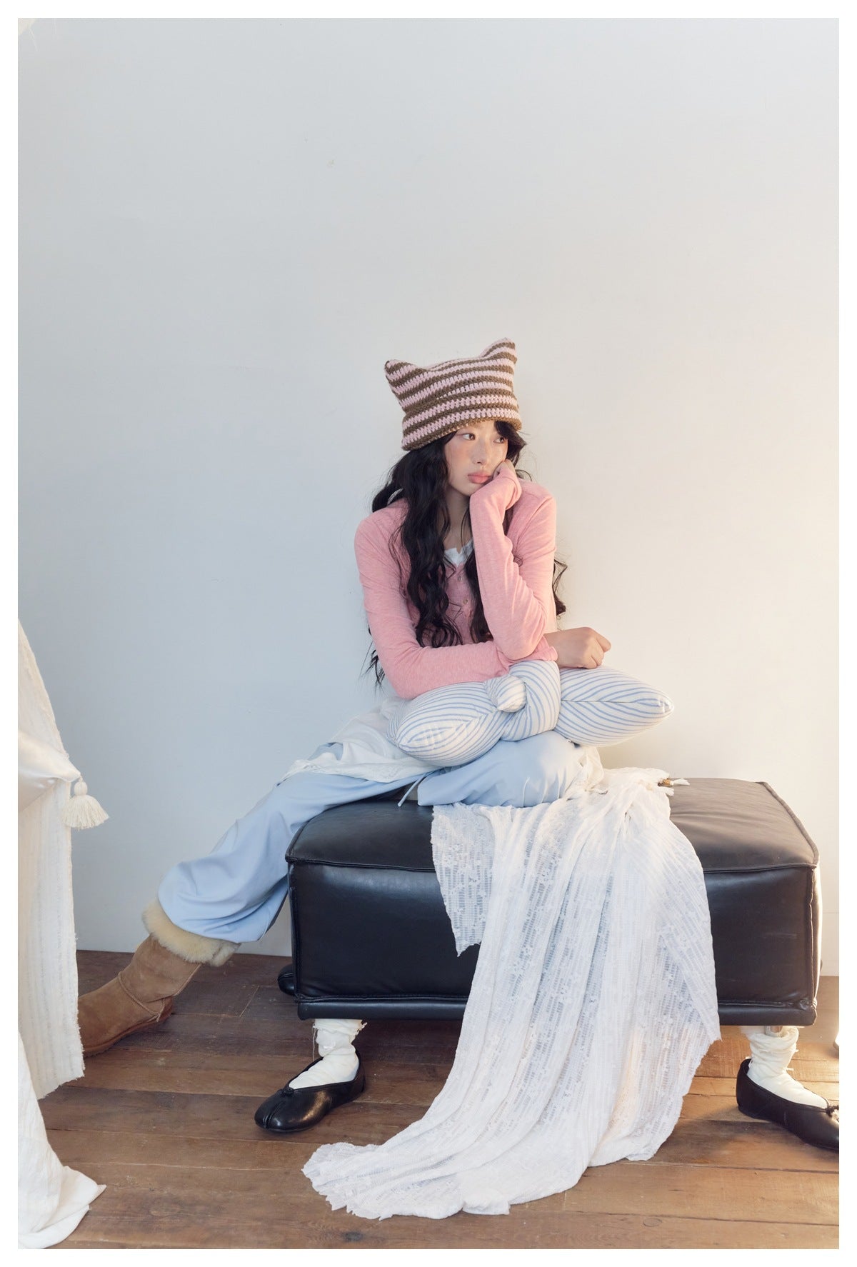 Seated model wears a pink long-sleeve henley, pale blue pants, striped leggings, and a brown-and-pink knit hat.