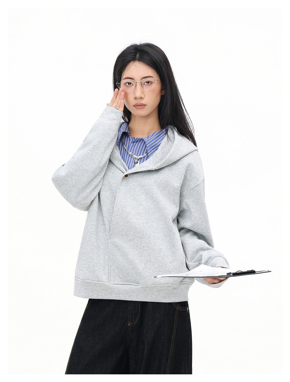 Model in a light gray hooded sweatshirt and dark jeans, raising one hand to her glasses while holding a clipboard.