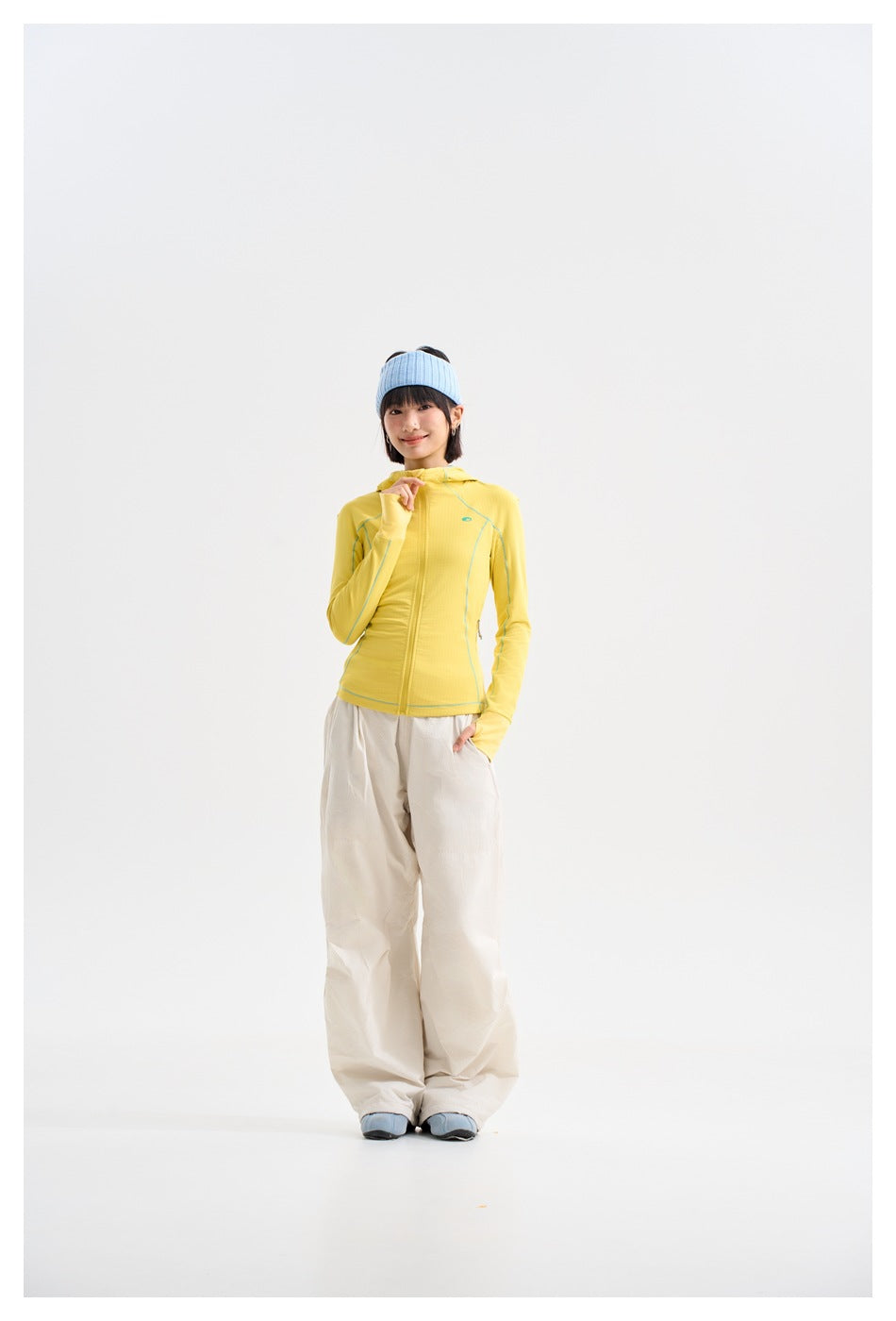 Model wearing a yellow zip-front hooded jacket with teal piping and loose cream pants, posing with a hand near the collar.