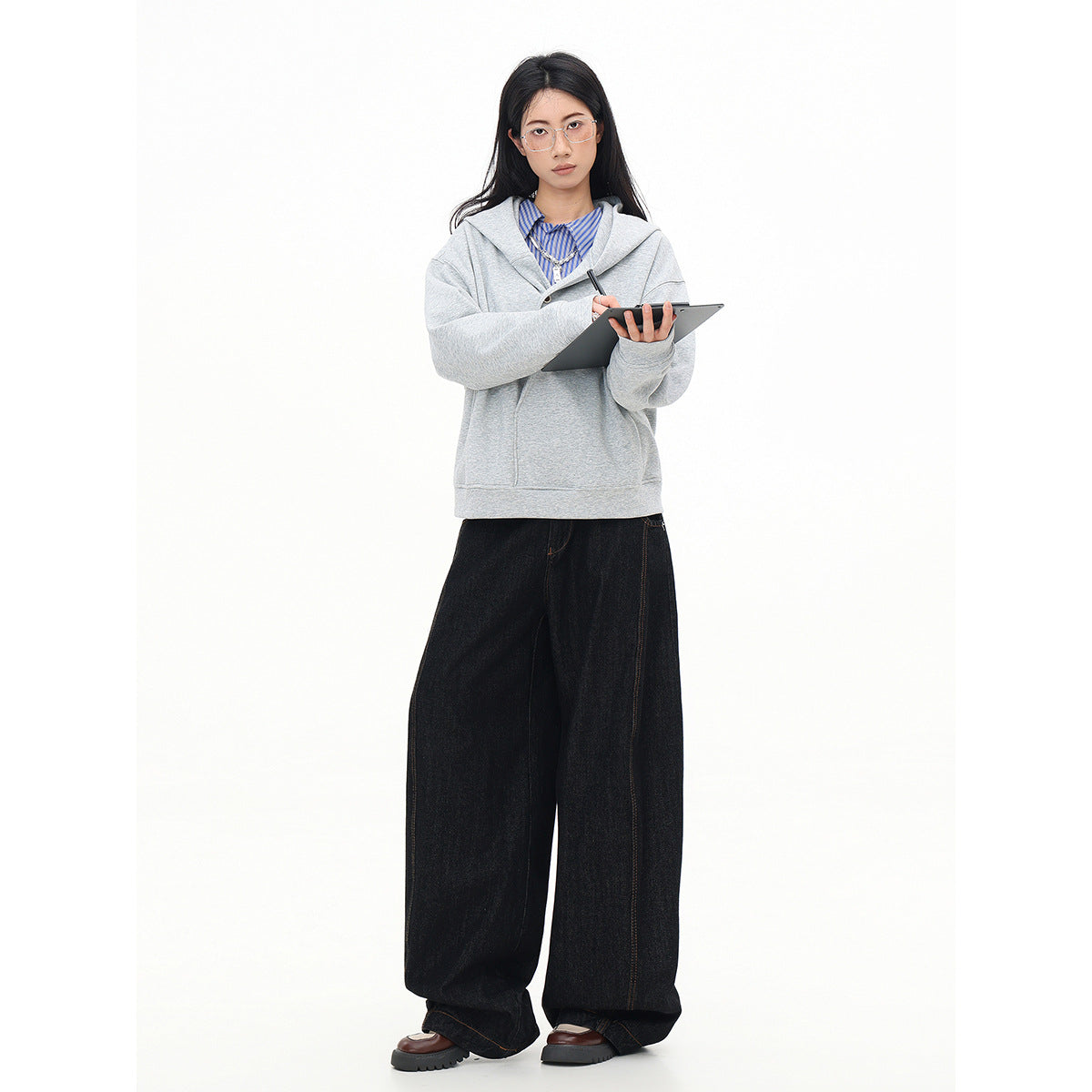 Full-length view of a model in a light gray hooded sweatshirt and dark wide-leg jeans, holding a tablet.