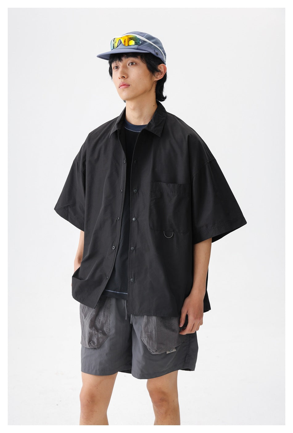 Model wearing an oversized black short-sleeve button-up shirt with a chest pocket and gray shorts against a white studio